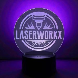 May include: A round, illuminated sign with the text "LASERWORKX" in white. The sign features a laser cutter graphic and a sunburst design. The base is black, and the background is a gradient of purple.