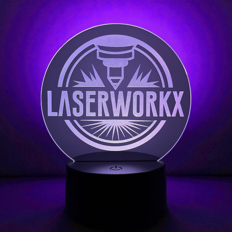 May include: A round, illuminated sign with the text "LASERWORKX" in white. The sign features a laser cutter graphic and a sunburst design. The base is black, and the background is a gradient of purple.