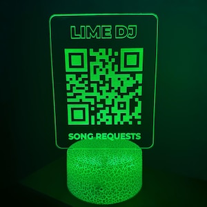 Op de afbeelding: Een groene LED-lamp met de tekst "LIME DJ" en een QR-code voor nummerverzoeken. De lamp staat op een getextureerde, cilindrische basis. Het ontwerp is modern en verlicht.