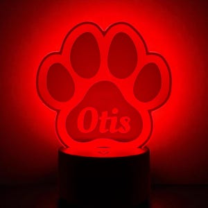 May include: A red LED night light shaped like a paw print, with the name "Otis" illuminated in the center. The light sits on a dark base, creating a glowing effect in a dim environment. Ideal for pet lovers.