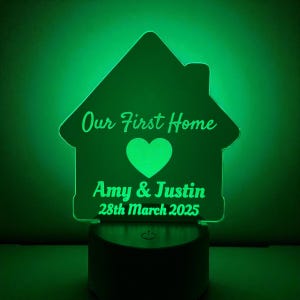 May include: A green-glowing house-shaped night light. The text "Our First Home" is above a heart, with "Amy & Justin" and the date "28th March 2025" below. The base is a dark, cylindrical shape.