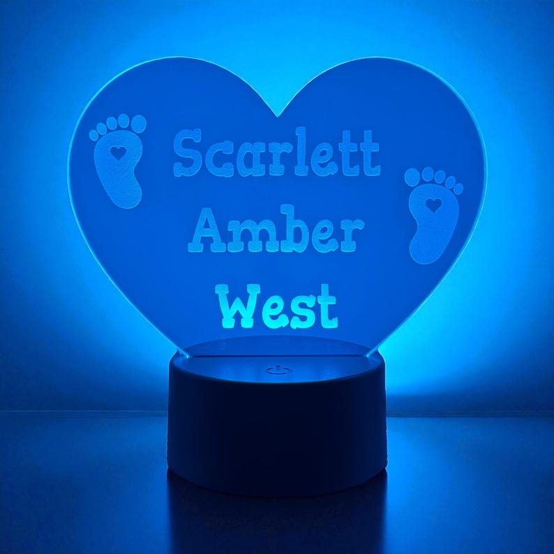 May include: A blue heart-shaped night light with the name "Scarlett Amber West" engraved on it. The light has two baby footprint designs. The base of the light is a dark color.