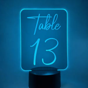 May include: A glowing blue LED table number sign. The sign displays the word "Table" in cursive script above the number "13". The base is black and round.