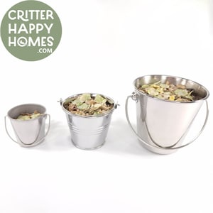 Può includere: Tre secchi di metallo argentato di diverse dimensioni riempiti con un mix di erbe e semi secchi. I secchi hanno manici e sono su uno sfondo bianco.