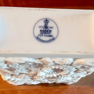 Puede incluir: Caja de baratijas de porcelana blanca con un dise&ntilde;o de relieve floral en la mitad inferior. La tapa presenta un emblema azul y blanco con el texto "KAISER GERMANY 125 JAHRE". La caja es rectangular con bordes redondeados.