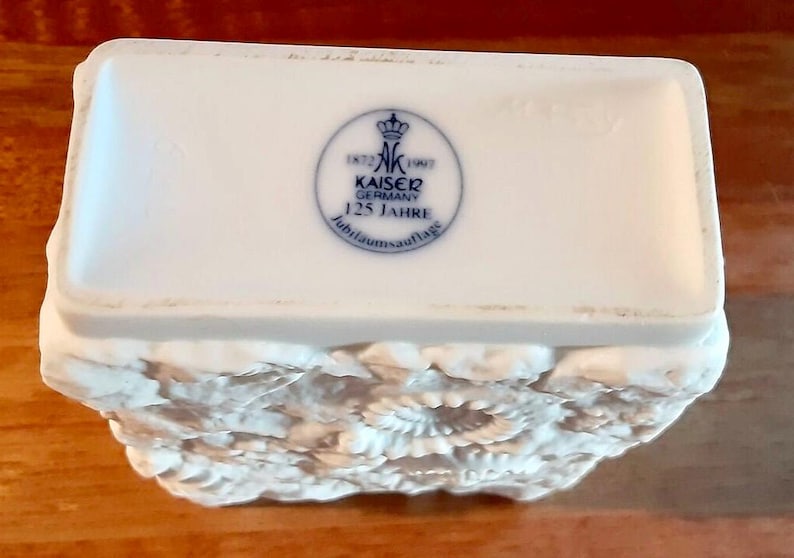 Puede incluir: Caja de baratijas de porcelana blanca con un dise&ntilde;o de relieve floral en la mitad inferior. La tapa presenta un emblema azul y blanco con el texto "KAISER GERMANY 125 JAHRE". La caja es rectangular con bordes redondeados.