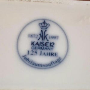 Puede incluir: Superficie de porcelana blanca con un emblema circular azul. El emblema presenta una corona, las letras "AK" y las palabras "KAISER GERMANY 125 JAHRE". Tambi&eacute;n son visibles los a&ntilde;os 1872 y 1997.