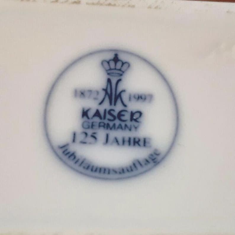Puede incluir: Superficie de porcelana blanca con un emblema circular azul. El emblema presenta una corona, las letras "AK" y las palabras "KAISER GERMANY 125 JAHRE". Tambi&eacute;n son visibles los a&ntilde;os 1872 y 1997.