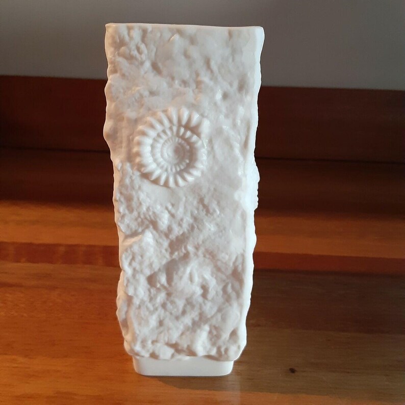Puede incluir: Un jarr&oacute;n rectangular blanco con una superficie texturizada similar a la piedra. El jarr&oacute;n presenta un dise&ntilde;o de f&oacute;sil en espiral en relieve. La base del jarr&oacute;n es blanca lisa y s&oacute;lida. El jarr&oacute;n mide aproximadamente 20 cm de alto.