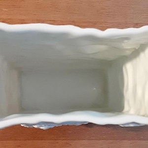 Puede incluir: Una jardinera rectangular de cer&aacute;mica blanquecina con un borde texturizado y ondulado. El interior es liso y de color claro. La jardinera est&aacute; dise&ntilde;ada para contener plantas y flores.