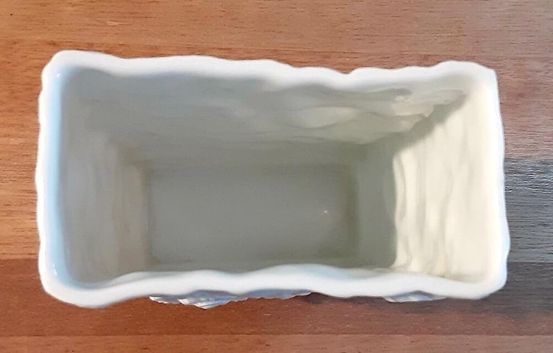 Puede incluir: Una jardinera rectangular de cer&aacute;mica blanquecina con un borde texturizado y ondulado. El interior es liso y de color claro. La jardinera est&aacute; dise&ntilde;ada para contener plantas y flores.
