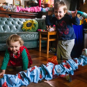 May include: Two young children playing with a colourful fabric tube in a living room. One child crawls on the floor, while the other holds the tube overhead. The room features a green sofa and wooden floors.