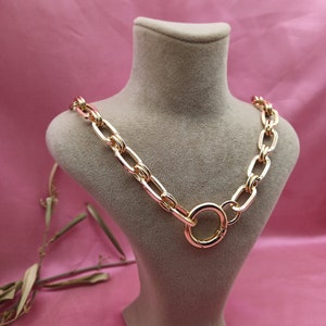 Könnte beinhalten: Eine goldene Kettenkette mit einem großen, runden, goldenen Verschluss. Die Kette befindet sich auf dem Hals einer Puppe.