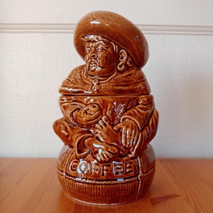 May include: A brown ceramic coffee jar shaped like a man wearing a wide-brimmed hat and sitting on a sack labelled "COFFEE".