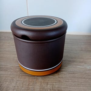 Könnte beinhalten: Ein brauner Keramiktopf mit Deckel. Der Topf hat eine zylindrische Form mit einem weißen und orangefarbenen Band am Boden. Der Deckel ist dunkelbraun mit einem helleren braunen und weißen Ringdesign. Der Topf steht auf einer Holzoberfläche.