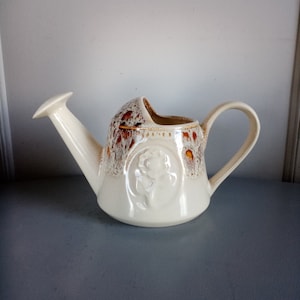 May include: A white ceramic watering can with a brown and orange glaze. The can has a spout and a handle. There is a raised design of a rose on the front of the can.
