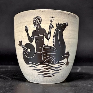Puede incluir: Una taza de cerámica color crema con una ilustración gráfica negra de una figura marina sosteniendo un tridente, montando una criatura con cola de aleta y cabeza de caballo, representada emergiendo de olas estilizadas en el frente.