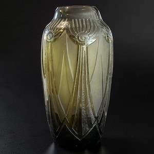 Può includere: Un vaso alto in vetro con una sfumatura di colore che va da una base verde oliva scuro a una parte superiore trasparente. L'esterno presenta un motivo a rilievo stampato con raggi geometrici verticali e motivi circolari con dettagli satinati.