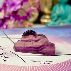 May include: A small, purple cloud-shaped crystal sits atop a book with the words "THE BOOK" printed on the cover. The crystal has a layered appearance with darker purple hues. The book has a pink and purple design with gold accents.