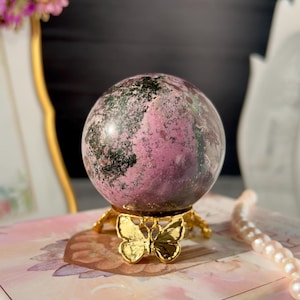 Puede incluir: Una esfera de cristal rosa y verde exhibida en un soporte dorado con forma de mariposa. La esfera tiene un aspecto veteado con tonos verdes oscuros y rosas. El soporte es ornamentado, con un diseño de mariposa en la base.