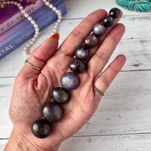 Puede incluir: Una mano que sostiene una fila de piedras preciosas pulidas de color oscuro. Las piedras varían en tonos de púrpura, gris y negro, con un acabado brillante. El fondo incluye libros, flores y objetos decorativos.