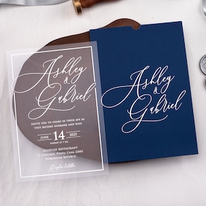 May include: A clear acrylic wedding invitation with white calligraphy that reads "Ashly & Gabriel" and details for the wedding ceremony. The invitation is on a wooden surface with a blue envelope.