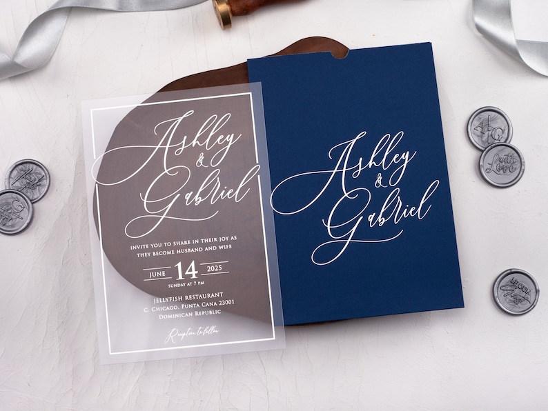 May include: A clear acrylic wedding invitation with white calligraphy that reads "Ashly & Gabriel" and details for the wedding ceremony. The invitation is on a wooden surface with a blue envelope.