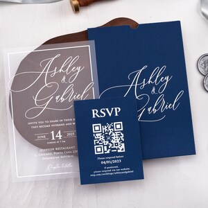 May include: A clear acrylic wedding invitation with a navy blue envelope. The invitation features the names "Ashley & Gabriel" in white calligraphy. The envelope also features the names "Ashley & Gabriel" in white calligraphy. The invitation includes the date, time, and location of the wedding. A separate RSVP card with a QR code is included.