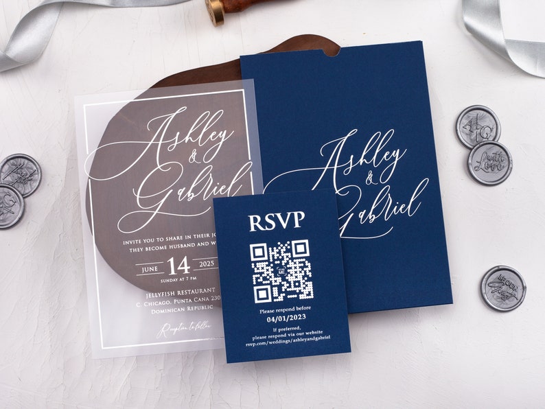 May include: A clear acrylic wedding invitation with a navy blue envelope. The invitation features the names "Ashley & Gabriel" in white calligraphy. The envelope also features the names "Ashley & Gabriel" in white calligraphy. The invitation includes the date, time, and location of the wedding. A separate RSVP card with a QR code is included.