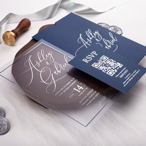 May include: A clear acrylic wedding invitation with a navy blue RSVP card. The invitation features the couple's names, "Ashley & Gabriel", in elegant calligraphy. The RSVP card includes a QR code for guests to respond.