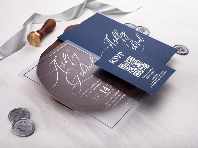 May include: A clear acrylic wedding invitation with a navy blue RSVP card. The invitation features the couple's names, "Ashley & Gabriel", in elegant calligraphy. The RSVP card includes a QR code for guests to respond.