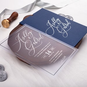 May include: A clear acrylic wedding invitation with a navy blue envelope. The invitation features the couple's names, "Ashley & Gabriel", in elegant white script. The text includes the date, time, and location of the wedding ceremony.