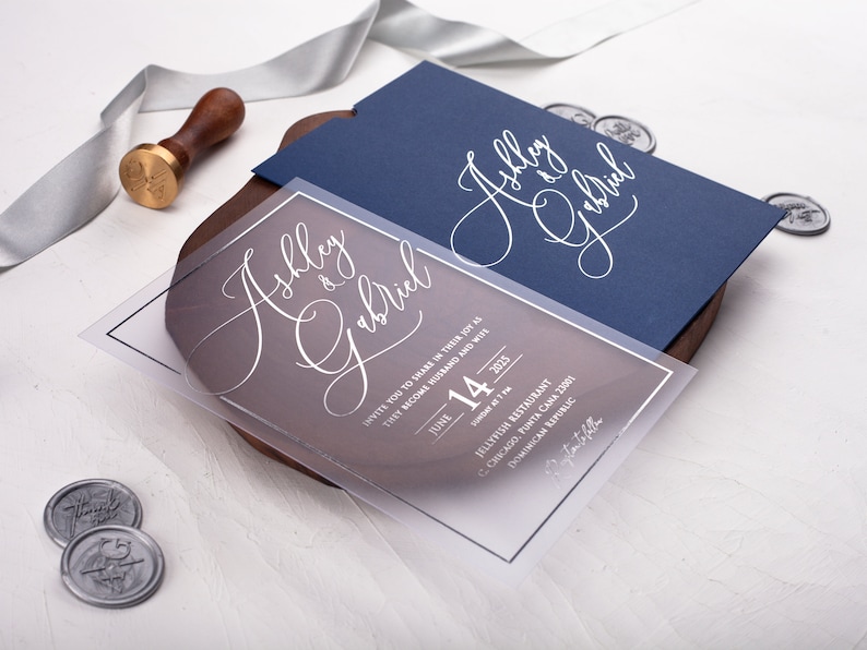 May include: A clear acrylic wedding invitation with a navy blue envelope. The invitation features the couple's names, "Ashley & Gabriel", in elegant white script. The text includes the date, time, and location of the wedding ceremony.