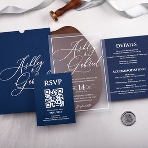 May include: A navy blue wedding invitation suite with a clear acrylic insert. The invitation features the couple's names, "Ashley & Gabriel", in white script. The RSVP card includes a QR code for guests to respond. The details card lists the ceremony location, date, and time, as well as accommodation options.