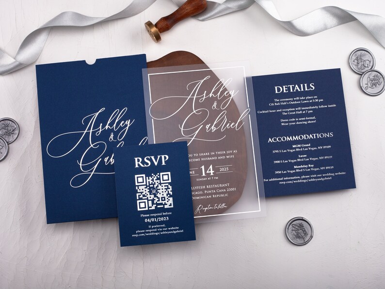 May include: A navy blue wedding invitation suite with a clear acrylic insert. The invitation features the couple's names, "Ashley & Gabriel", in white script. The RSVP card includes a QR code for guests to respond. The details card lists the ceremony location, date, and time, as well as accommodation options.