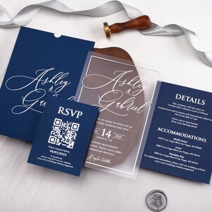 May include: A navy blue wedding invitation suite with a clear acrylic invitation card featuring the names "Ashley & Gabriel" in white cursive script. The RSVP card has a QR code and the text "Please respond by 04/01/2023". The details card has the text "DETAILS" and "ACCOMMODATIONS" in white.