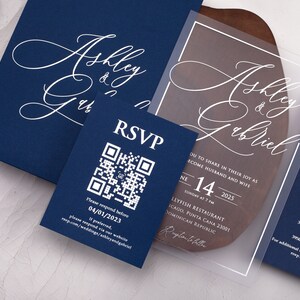 May include: A clear acrylic wedding invitation with white calligraphy text that reads "Ashley & Gabriel". The invitation includes the date, time, and location of the wedding. The invitation is on a wooden surface.