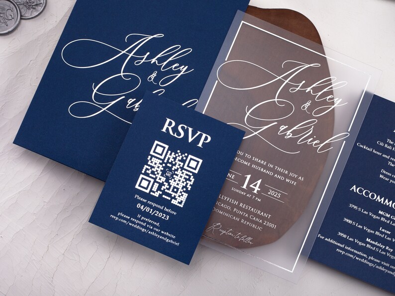 May include: A clear acrylic wedding invitation with white calligraphy text that reads "Ashley & Gabriel". The invitation includes the date, time, and location of the wedding. The invitation is on a wooden surface.