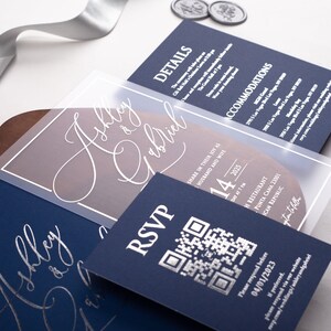 May include: A navy blue wedding invitation with a clear overlay featuring the names "Ashley & Gabriel" in white calligraphy. The overlay also includes a QR code and the date "14" in a large font. The text "RSVP" is printed in white on the navy blue card.