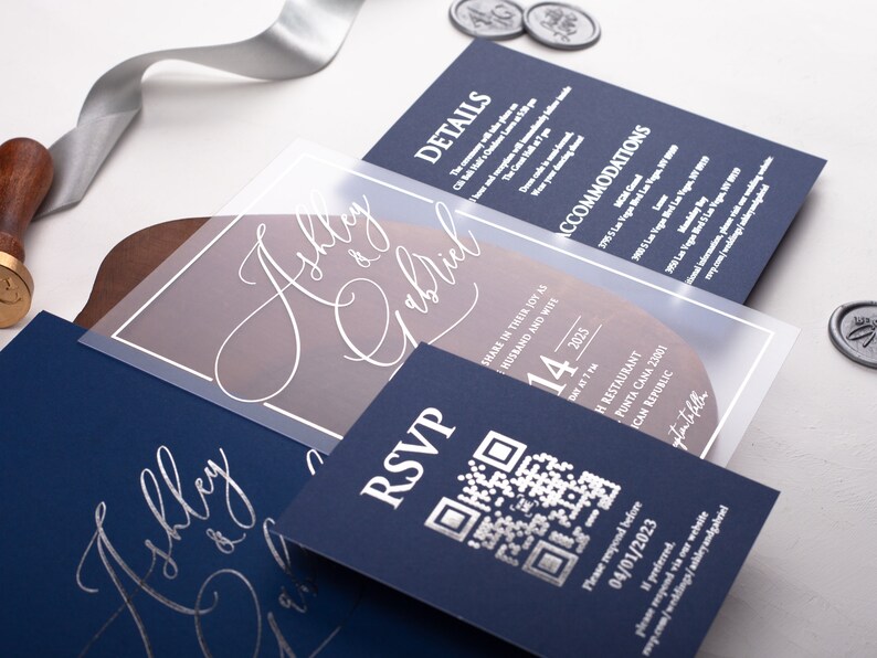May include: A navy blue wedding invitation with a clear overlay featuring the names "Ashley & Gabriel" in white calligraphy. The overlay also includes a QR code and the date "14" in a large font. The text "RSVP" is printed in white on the navy blue card.