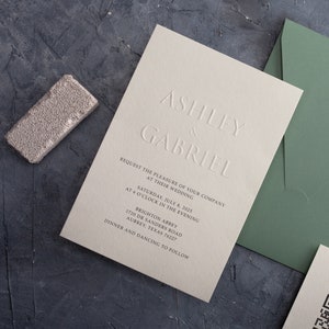 Puede incluir: Conjunto de invitaciones de boda con texto en relieve, incluyendo los nombres "Ashley & Gabriel" y detalles del evento. Incluye una invitación gris claro, un sobre verde y un objeto rectangular plateado texturizado. Sobre una superficie gris oscura.