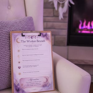 May include: A wooden clipboard holding a printed sheet titled The Wisdom Beneath, featuring purple watercolor accents, moon phases, and prompts for self-reflection. The page includes sections for writing, a central affirmation box, and small icons like a heart and lotus.