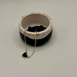 May include: A gold-colored chain necklace with a black heart-shaped pendant. The necklace is displayed near a small, round, decorative bowl with a white, black, and red speckled design.
