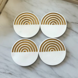 May include: Set of four white round coasters with a gold rainbow design. Each coaster has a half-circle of gold rainbow design and a half-circle of white.