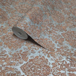 May include: A roll of wallpaper with a detailed damask pattern in copper and gray. The design features swirling floral and leaf motifs, creating a textured, elegant look. The wallpaper is partially unrolled, revealing the pattern.