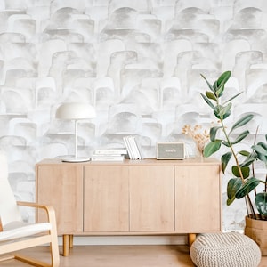 May include: A light wood cabinet with three doors and a white lamp on top. The cabinet is in front of a wall with a gray and white abstract pattern wallpaper. A small speaker and a stack of books are on the cabinet. A potted plant is on the right side of the cabinet.