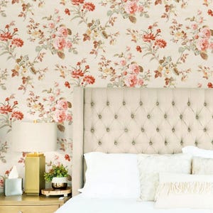 May include: A bedroom scene featuring a tufted headboard, white pillows, and a floral wallpaper with red and pink roses. A gold lamp and a small plant sit on a wooden nightstand. The bed is made with white sheets.