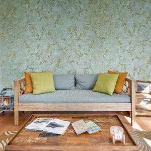 May include: A light blue sofa with wooden arms and legs, two orange pillows, and one green pillow. The sofa is in front of a wall with a green and gold textured wallpaper. There are two books and a candle on a wooden coffee table in front of the sofa.