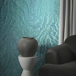 May include: A dark gray ceramic vase sits atop a white pedestal against a teal and blue patterned wall. A dark gray armchair and white curtains are in the background. The wall has a flowing, marbled design.