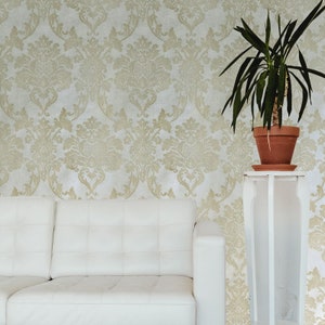 May include: A white couch with a white throw pillow sits in front of a wall with a gold damask pattern. A potted plant sits on a white stand to the right of the couch.